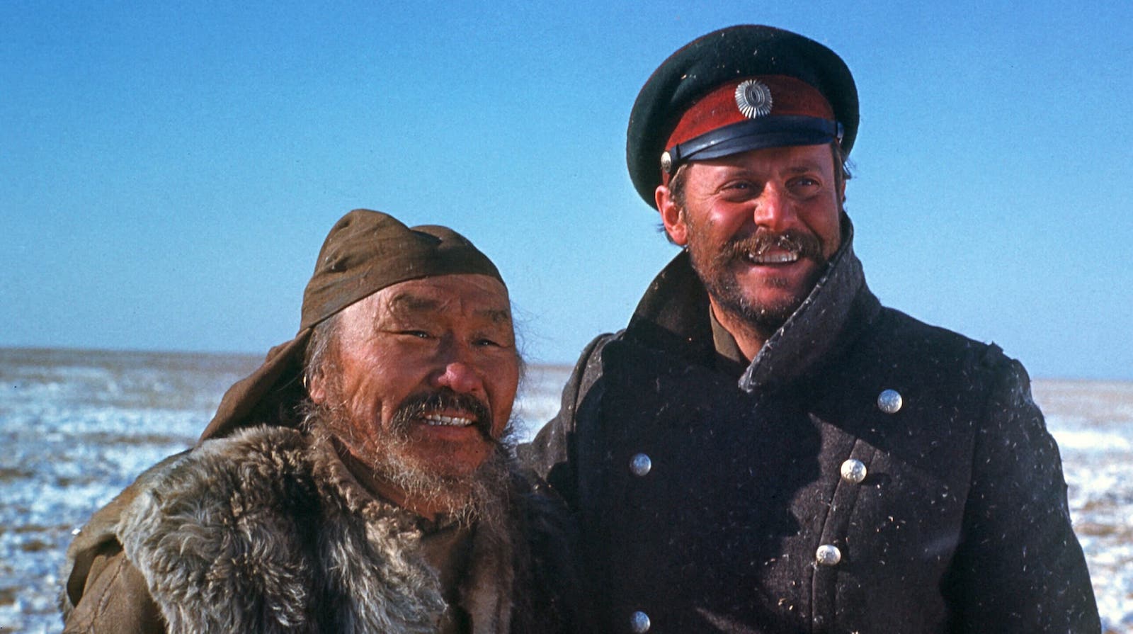 Two men smile against a snowy landscape and a bright blue sky.