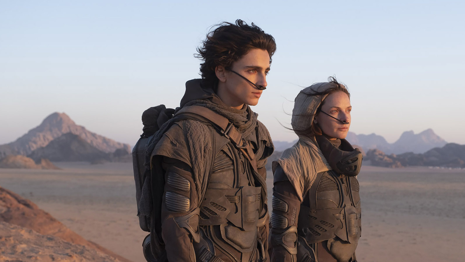 A young man and woman stand in a desert with futuristic beige costumes and breathing apparatuses connected to their noses.