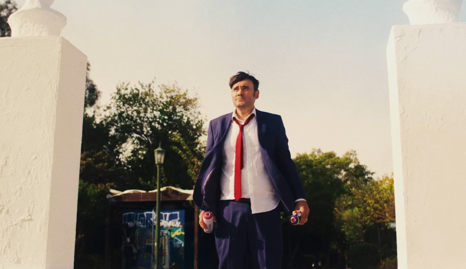 A man in a rumpled blue shit and red tie with loose collar stands tensely outside in front of trees in a public park with a pained expression.