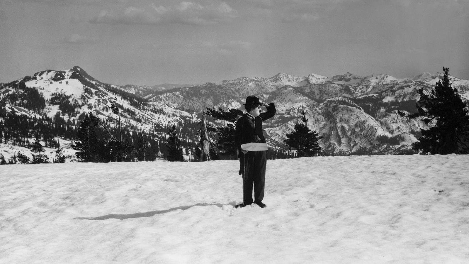 A man dressed in a bowler hat and a little tramp outfit stands on a snowy mountainside his hand above his forehead as he looks at the landscape.