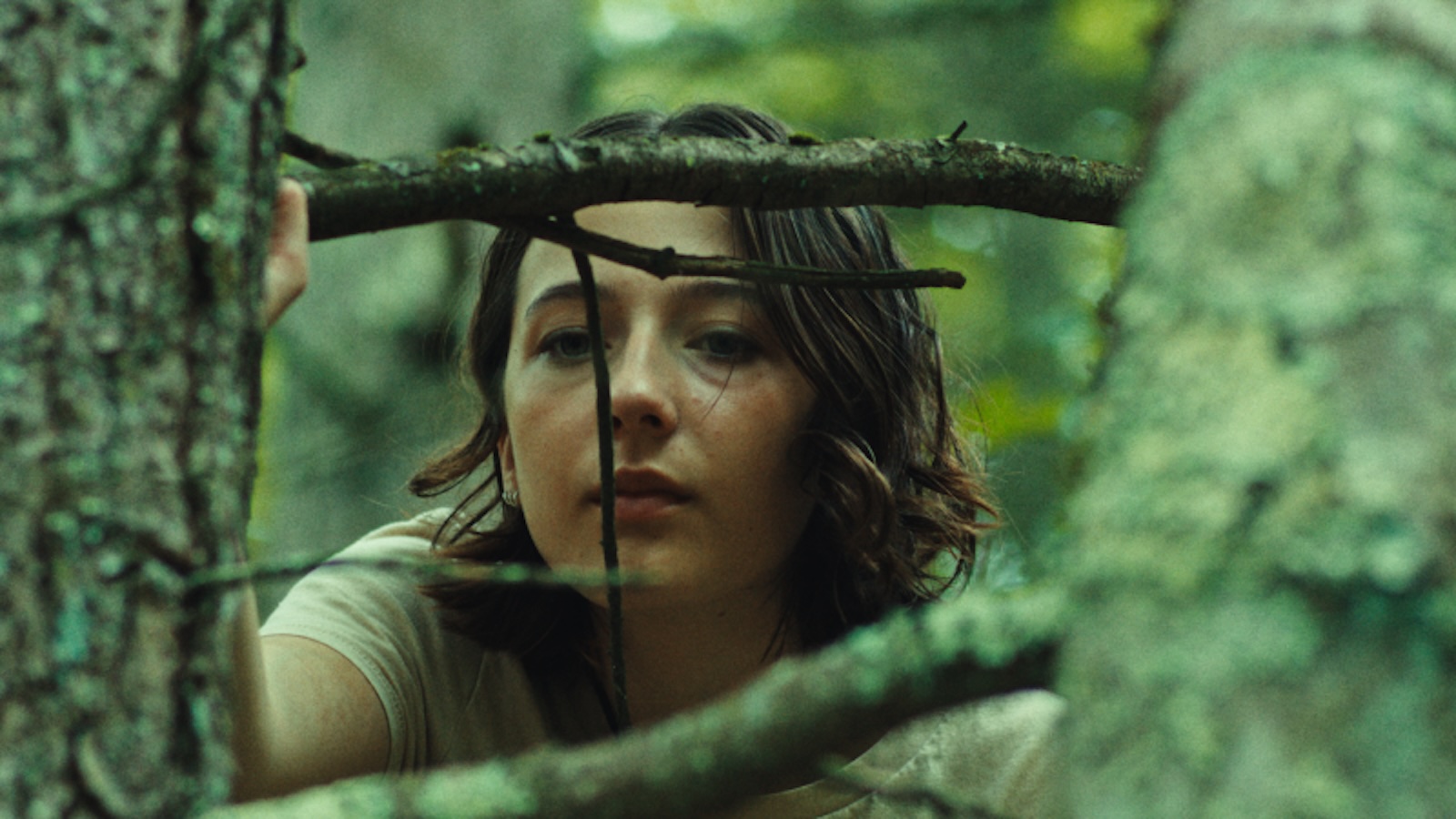 A teenage girl with chin-length brown hair stands in a green-tinted forest looking through branches.