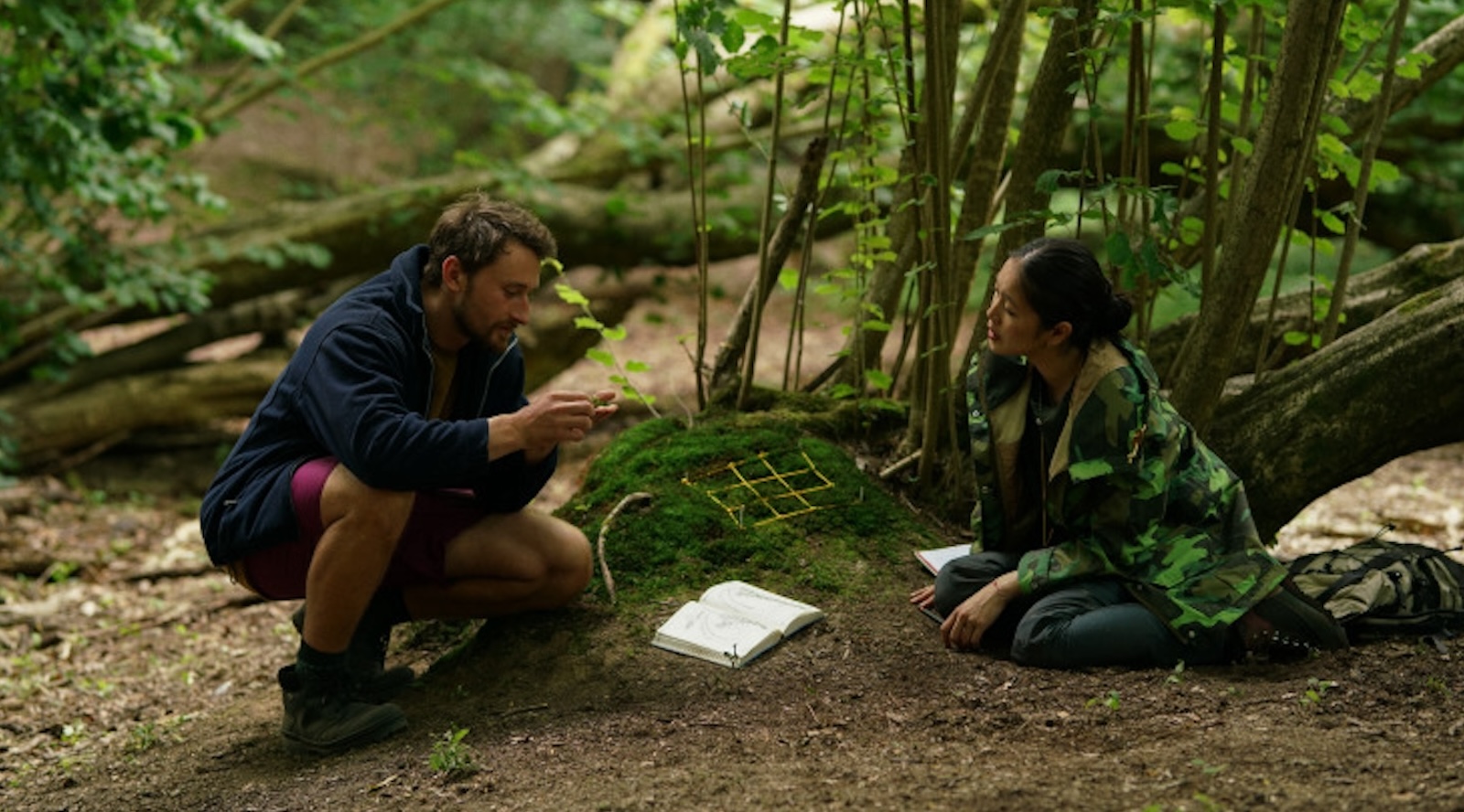 A man in shorts and a blue jacket kneels in front of a woman sitting in a green coat. They are in a forest, with an open book lying on the ground between them.