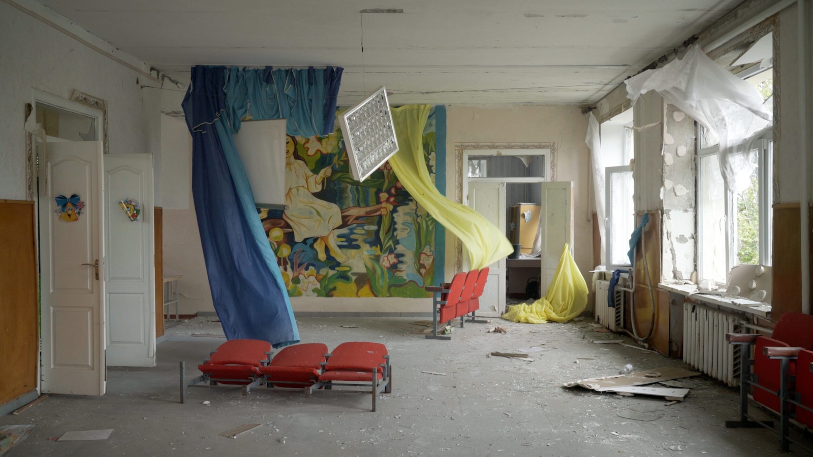 An abandoned room that appears to be in the aftermath of some destruction, with yellow and blue curtains fluttering and red chairs strewn about the room. The windows on the right are missing and appear to have been blown out.