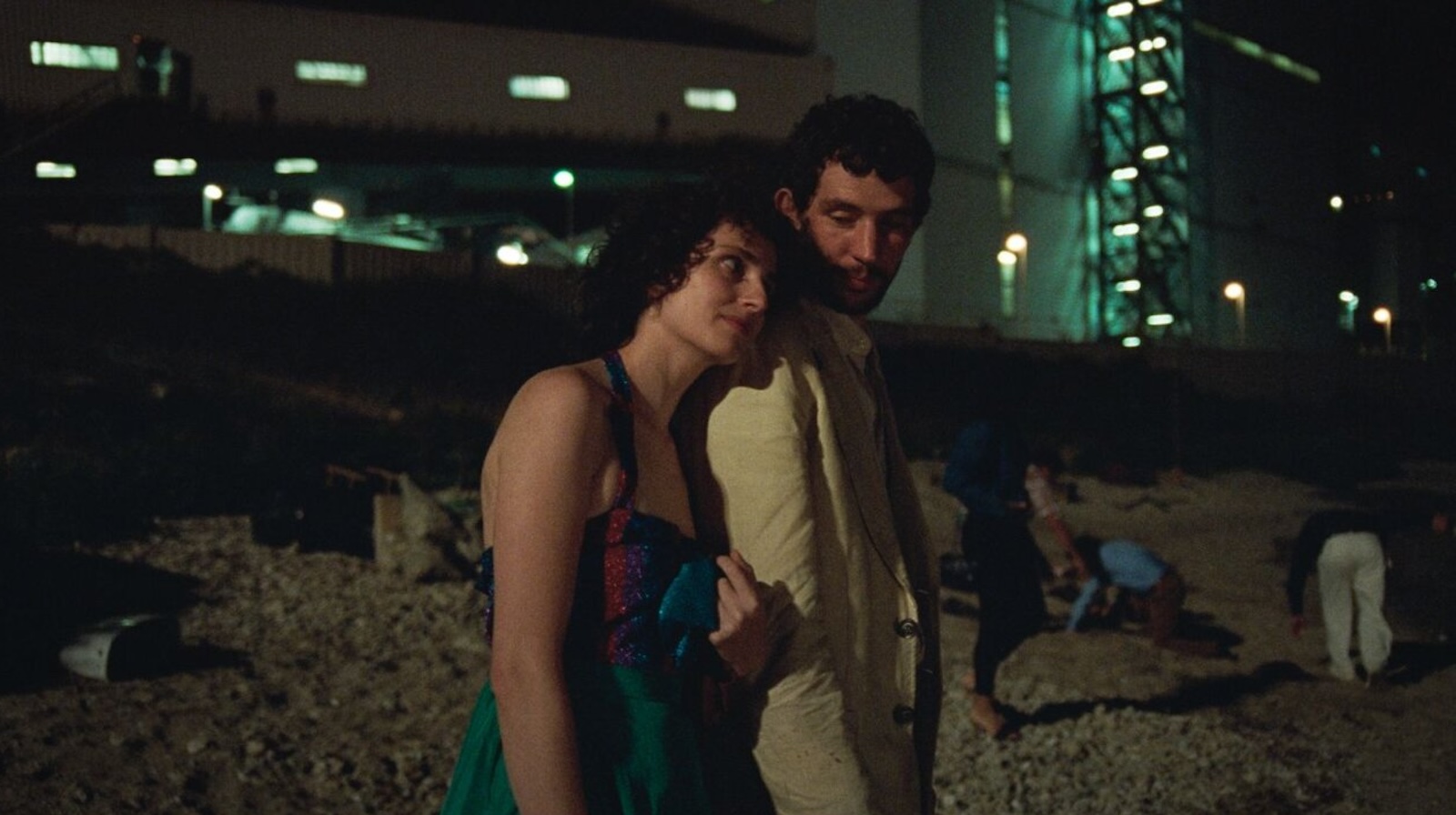 An unshaven man in a rumpled beige suit embraced by a woman in a green dress outside at an excavation site at night
