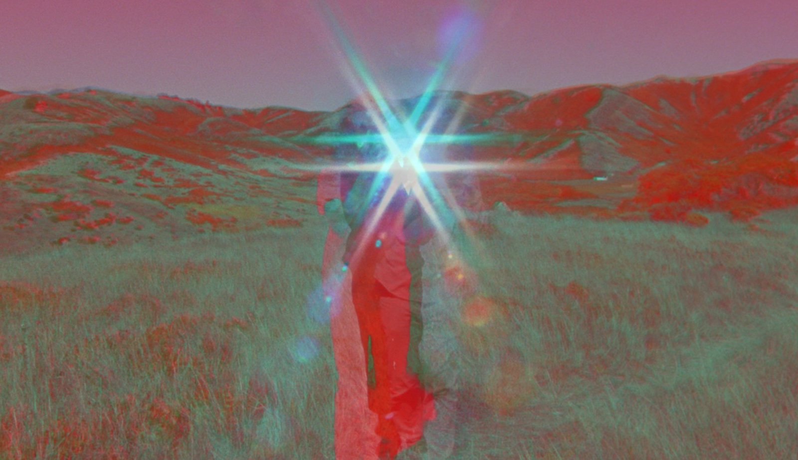 A blurry, surreal image of red and green of a figure in a red dress standing in a field with a glowing light in the middle of the frame.