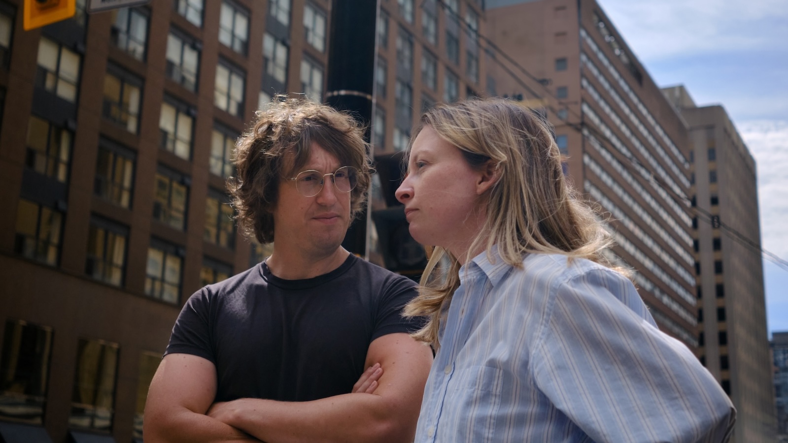 A man with glasses and shaggy brown hair and a woman with long tousled blonde hair stand next to each other at a low angle against a city streetscape with tall buildings. He looks at her, she looks away.