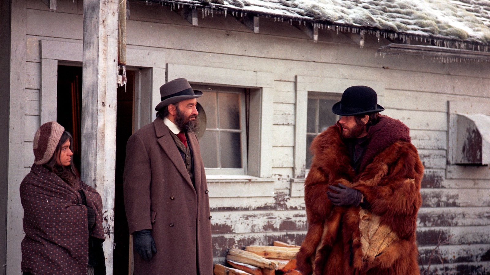 A bearded man in a bowler hat and enormous fur coat stands outside in a snowy old western town talking to a man and woman.