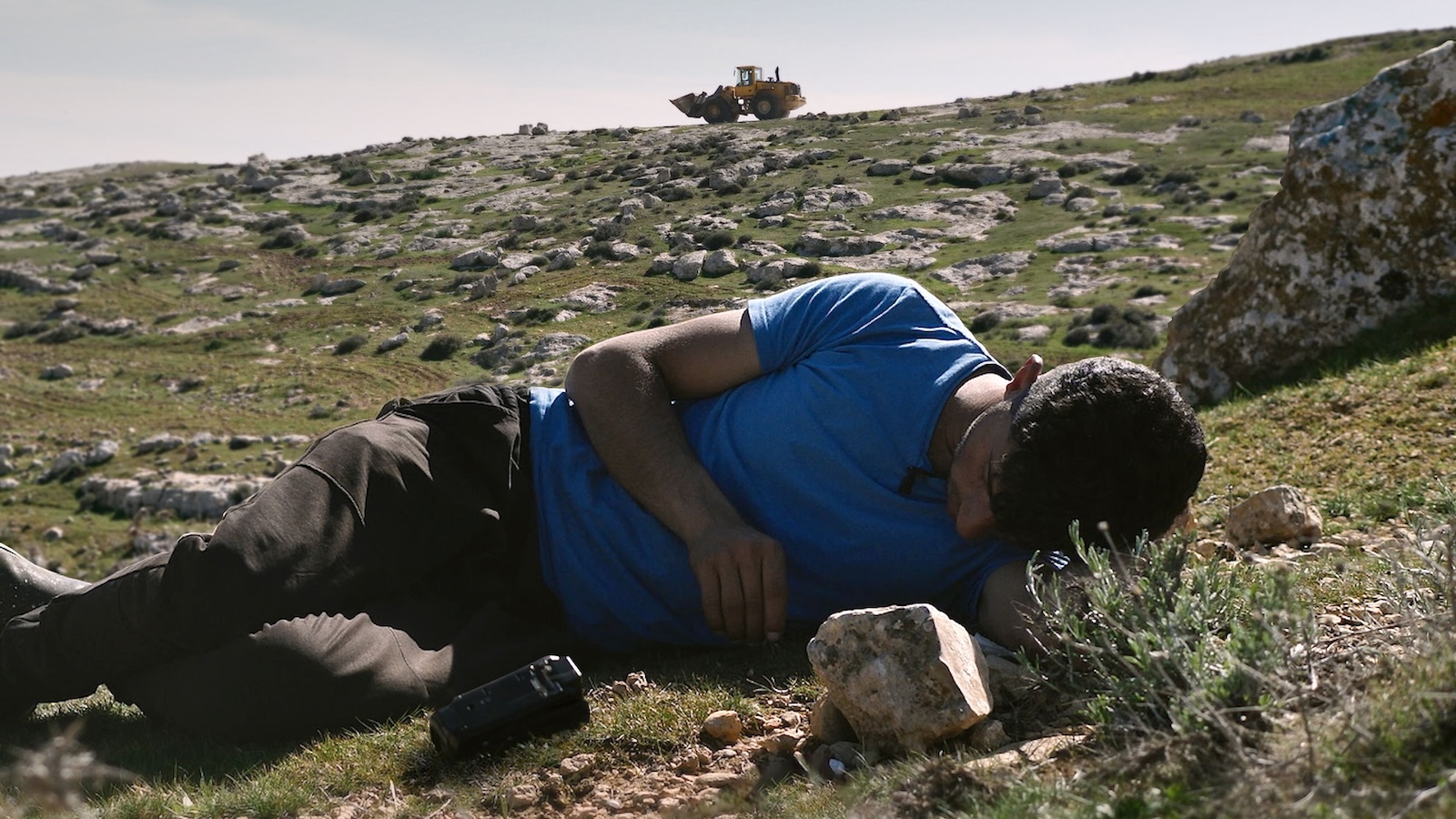A man in a blue t-shirt reclines in a rocky and green landscape looking at the ground. In the far distance bulldozer can be seen on the horizon.