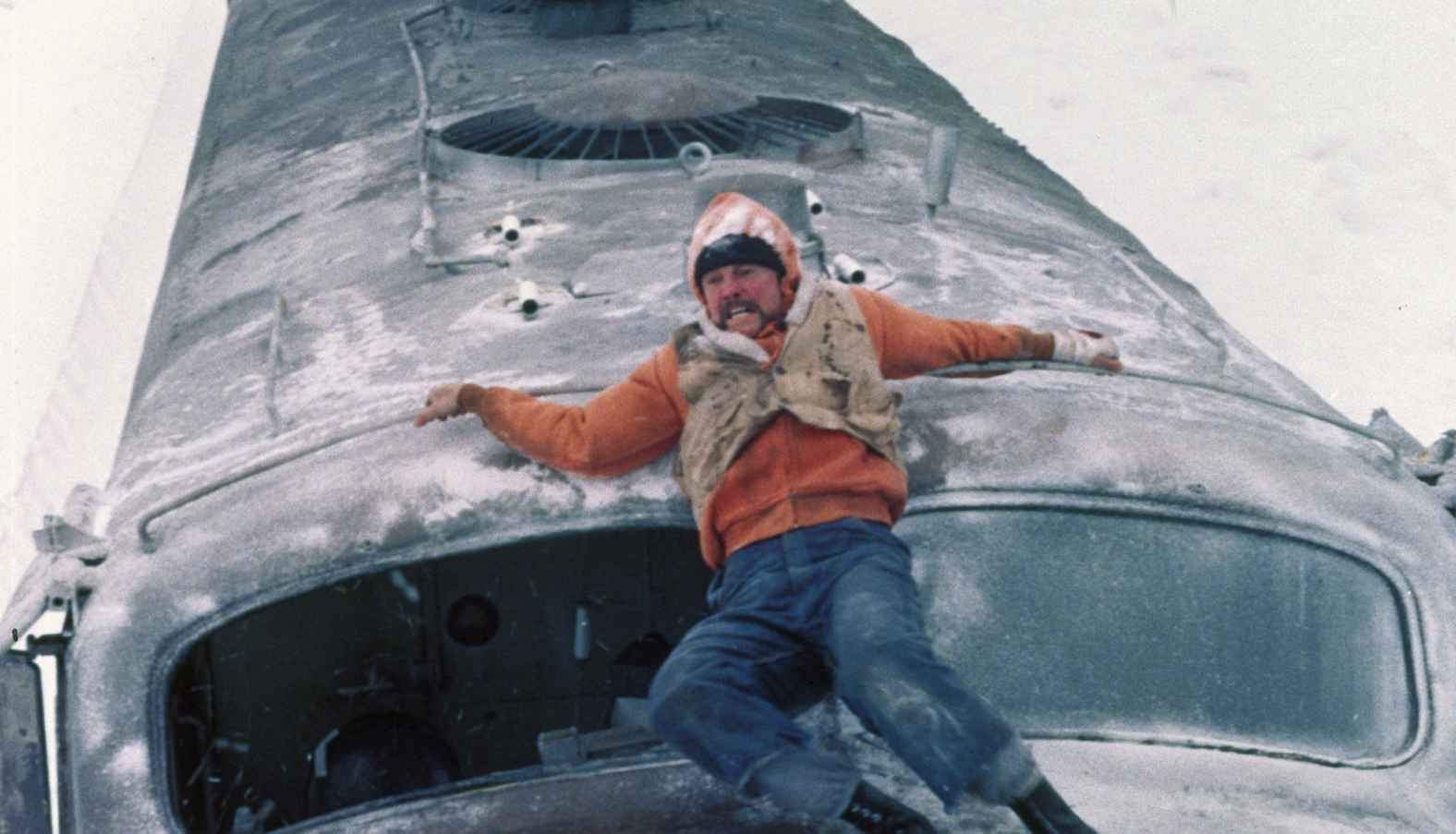 A man in an orange shirt and beige vest desperately holds to the top of a train as it runs through a snowy landscape.