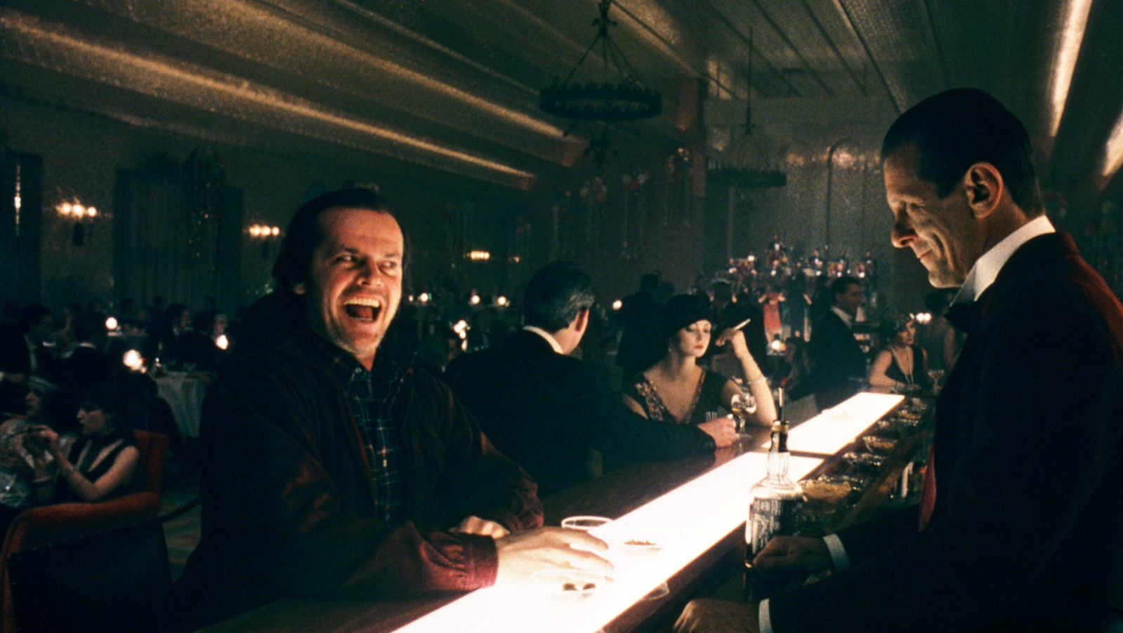 A man with a maniacal smiling face sits at a bar with light emanating from below, with a bartender looking at him eerily.