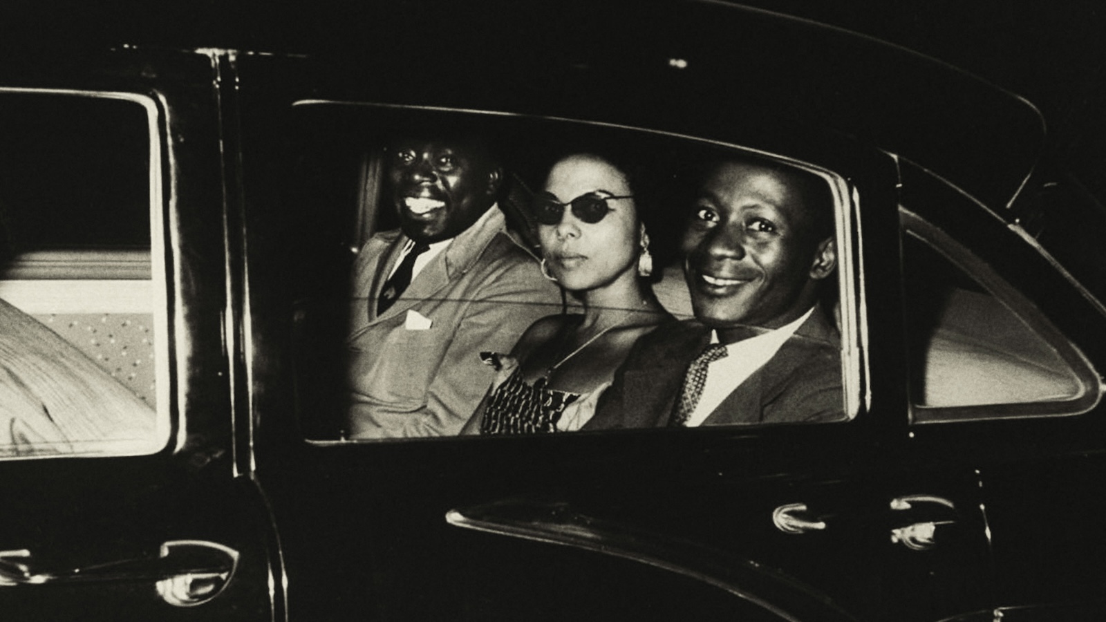 A black and white photo from the sixties of three people in the back of a town car, looking at camera, two dark-skinned Black men and a lighter-skinned Black woman between them; the men smile, the woman purses her lips.