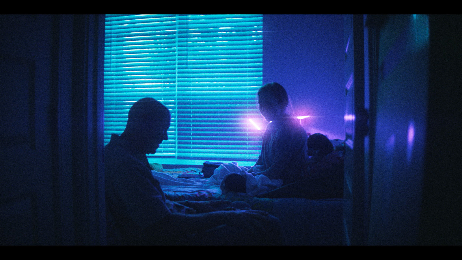 A young female figure sits cross-legged on a bed in a dark room with the silhouette of an older bald man sitting near her in a chair, as blue light streams through a window with half-open blinds.