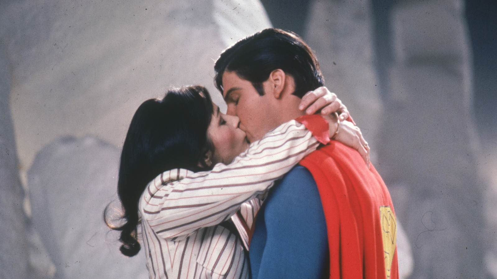 Superman, wearing a red cape and blue suit, kisses a dark-haired woman, Lois Lane, in a striped blazer.