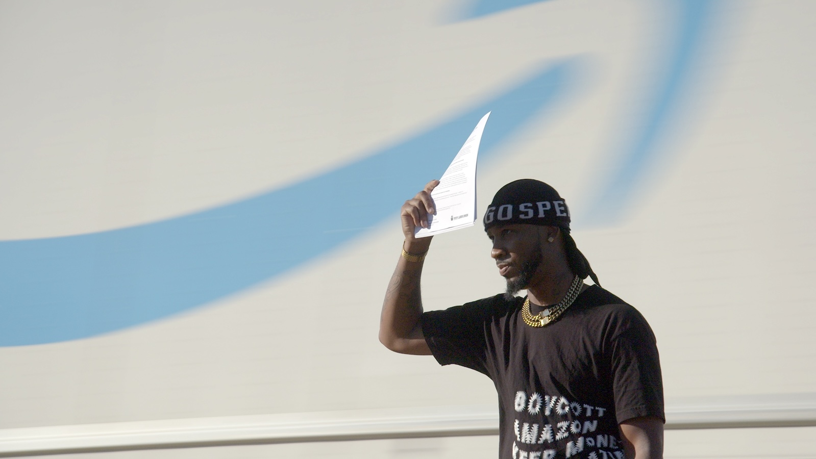 A man wearing a knit cap and a T-shirt that says "BOYCOT AMAZON" holds a piece of paper over his head against the side of a white truck with blue lettering.