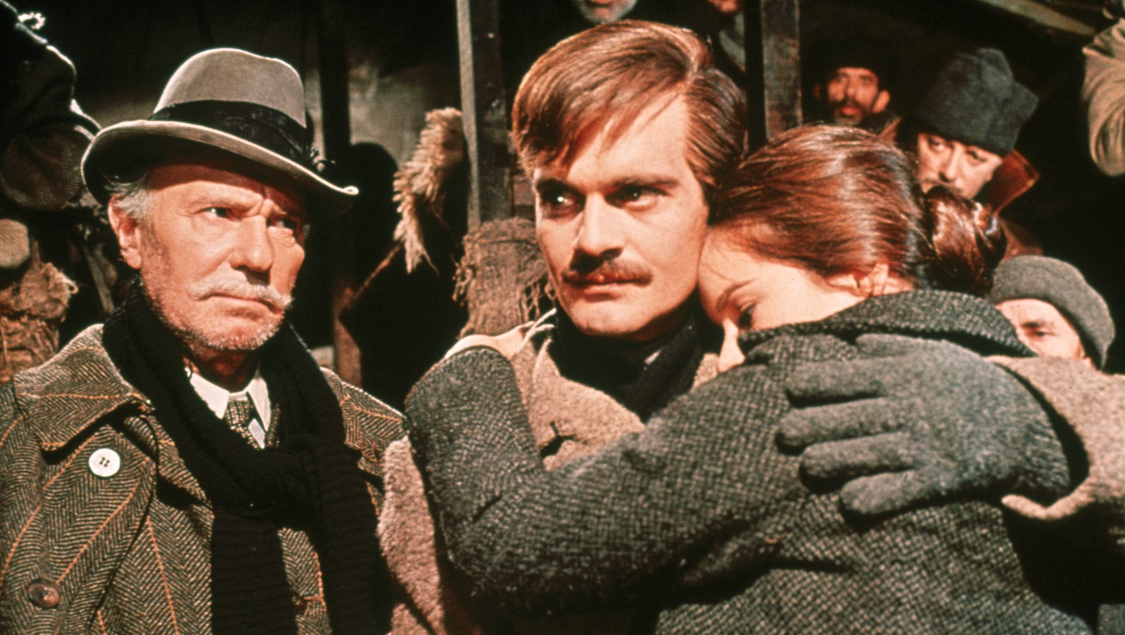 A mustachioed man embraces a woman, both are wearing thick winter clothes, and a white-haired man with a white mustache watches them.