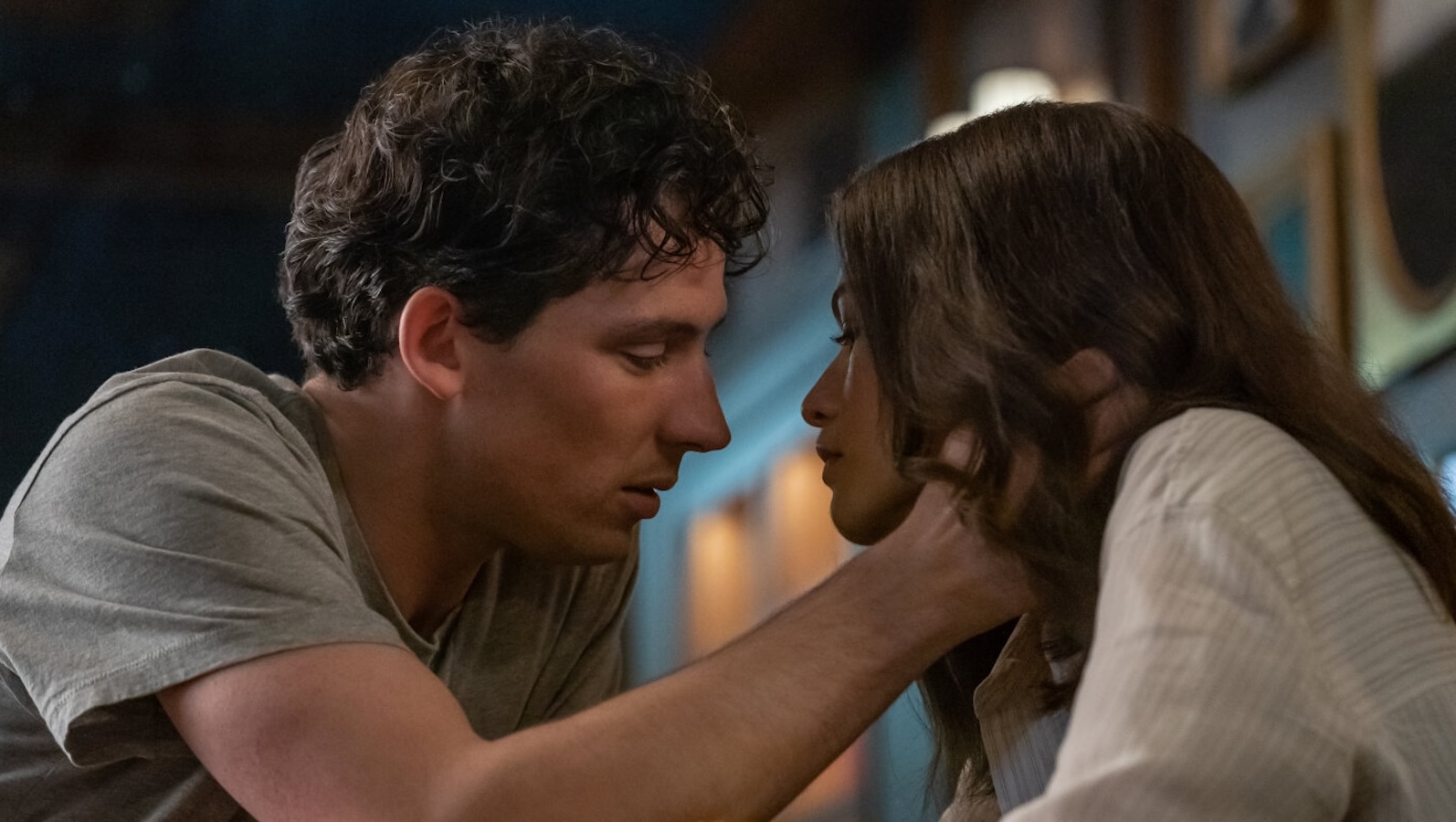 A young man with tousled dark hair reaches for a young woman with long dark hair and embraces her cheek as he reaches in for a kiss