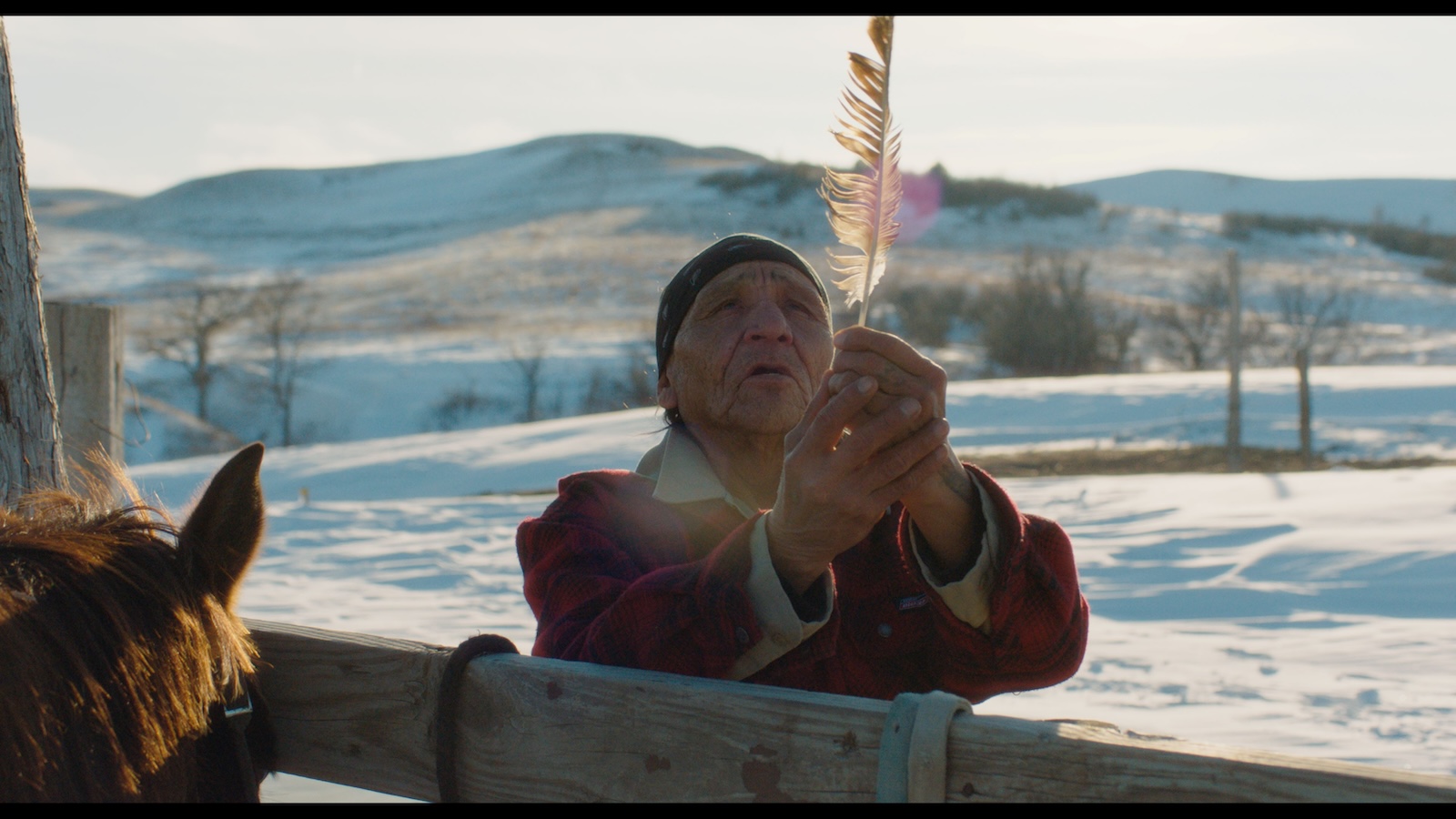 A Native American man prays to the sky holding a feather against a snowy country backdrop, standing next to a fence. Next to the fence there is the top of a brown horse's head.