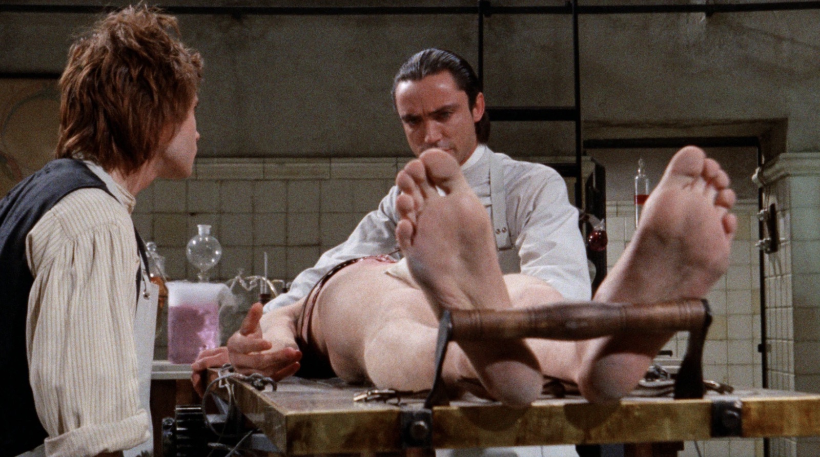 A mad scientist with dark slicked back hair and his redheaded male assistant stand over a naked body buckled to an operating table, the bottoms of its feet facing us.
