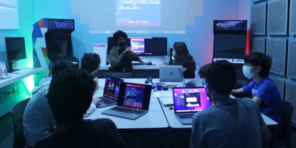 A group of young people sit at tables looking at laptops working together on coding.