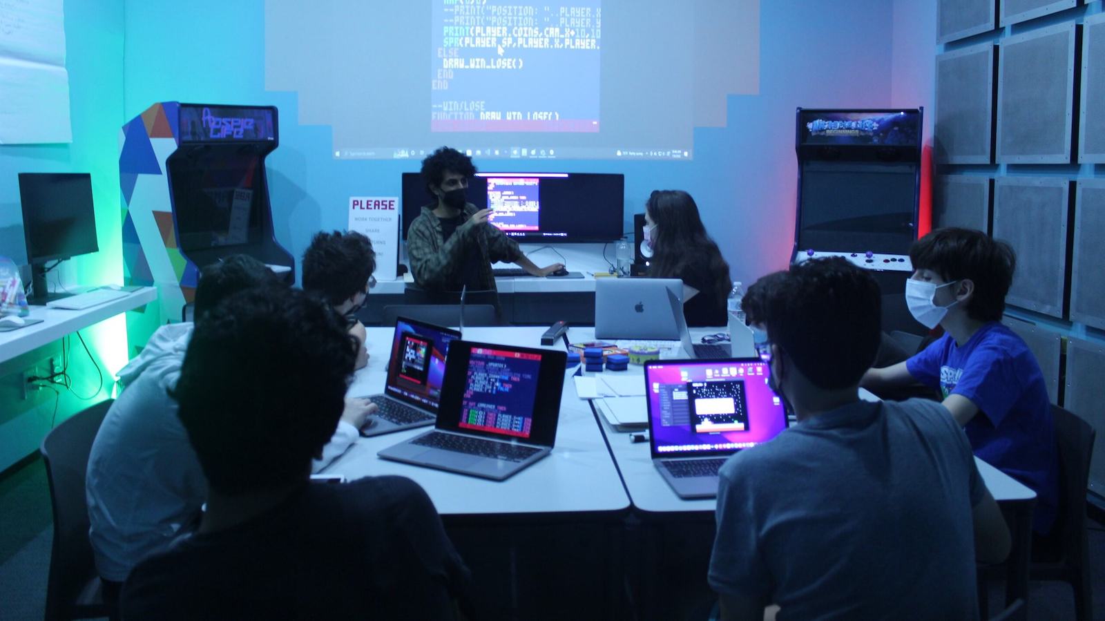 A group of young people sit at tables looking at laptops working together on coding.