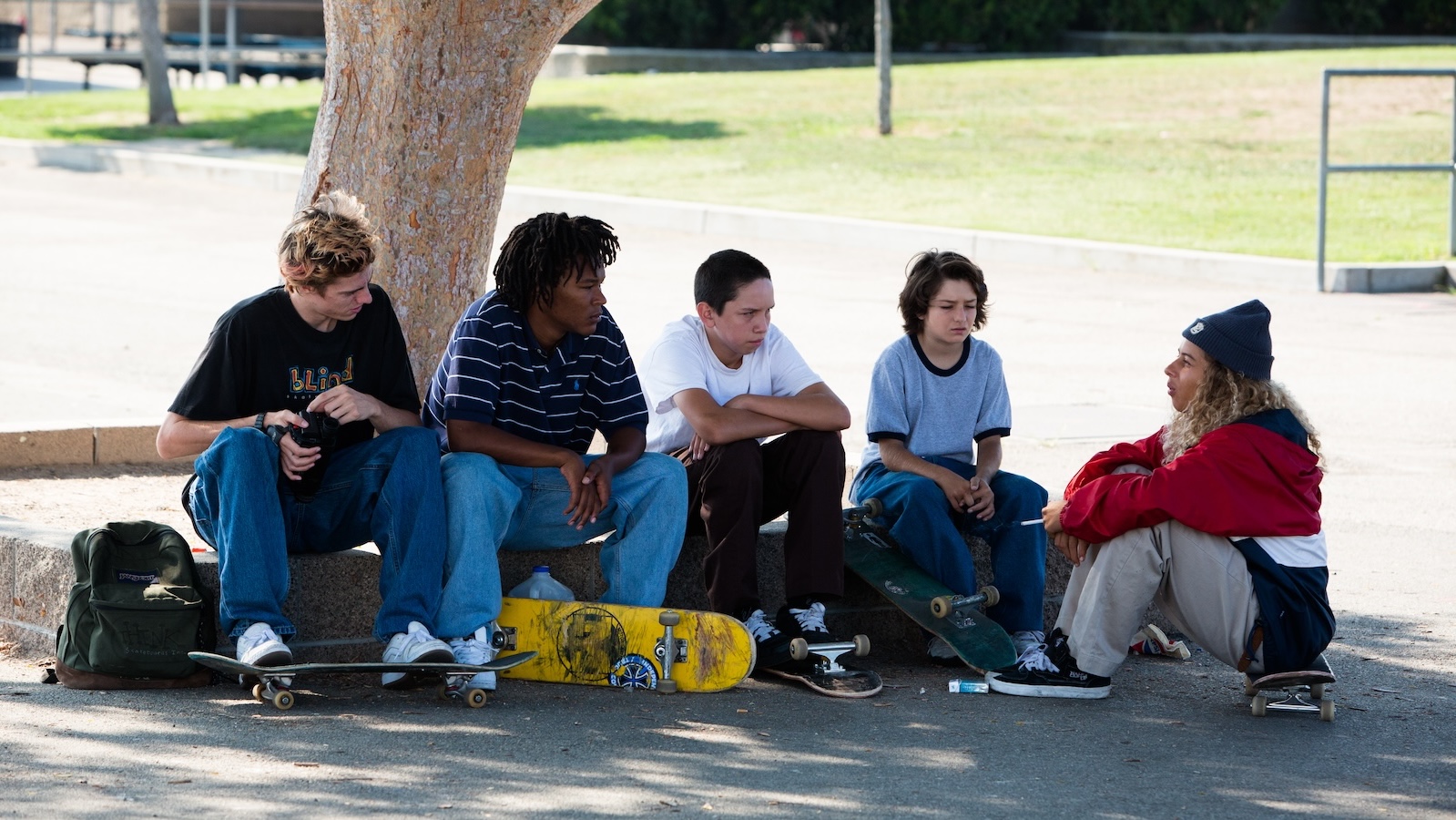 A line of five kids either sitting on or putting their feet on their skateboards sit on a suburban street on a sunny street in the afternoon