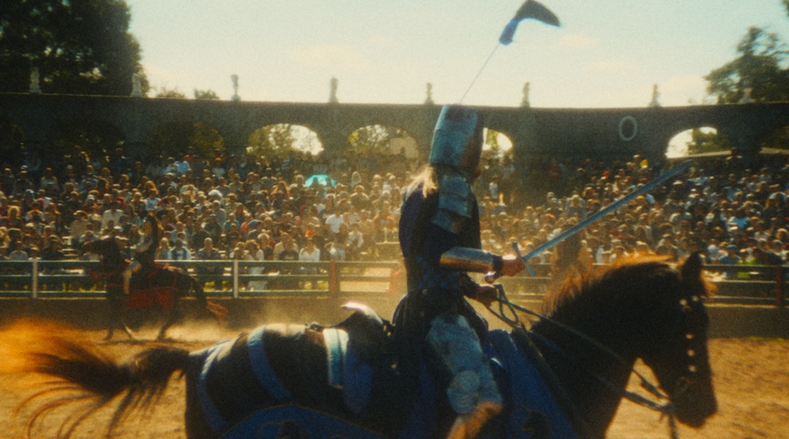 An armored knight rides on a horse in the middle of a Renniassance faire grounds, sword raised, looking at bleachers full of people watching.