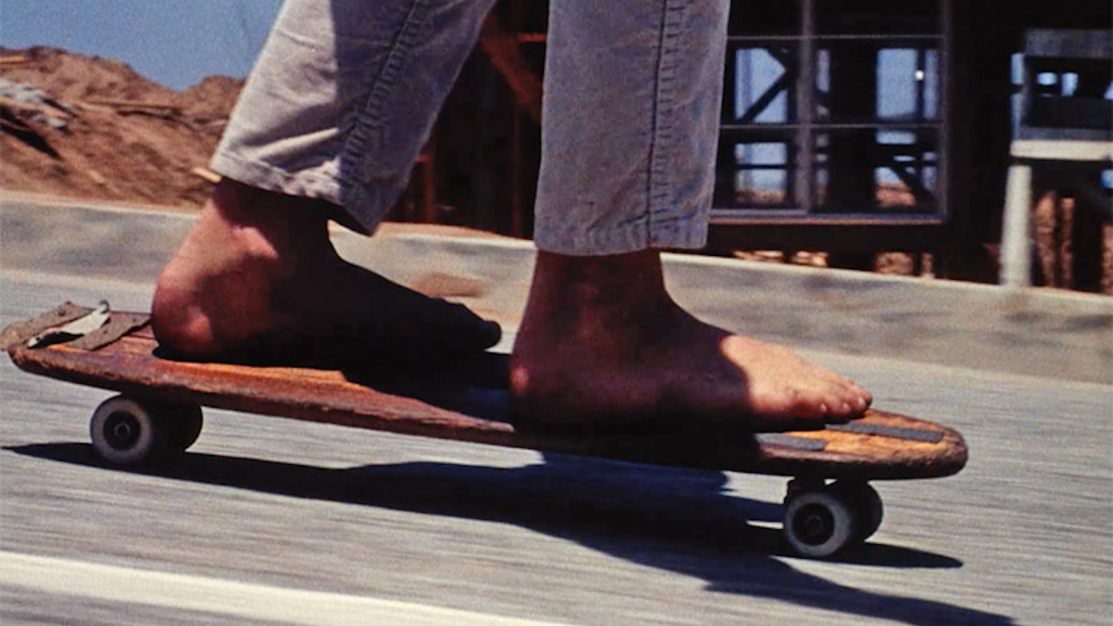 A close up of two bare feet on a skateboard as it rolls downhill on pavement