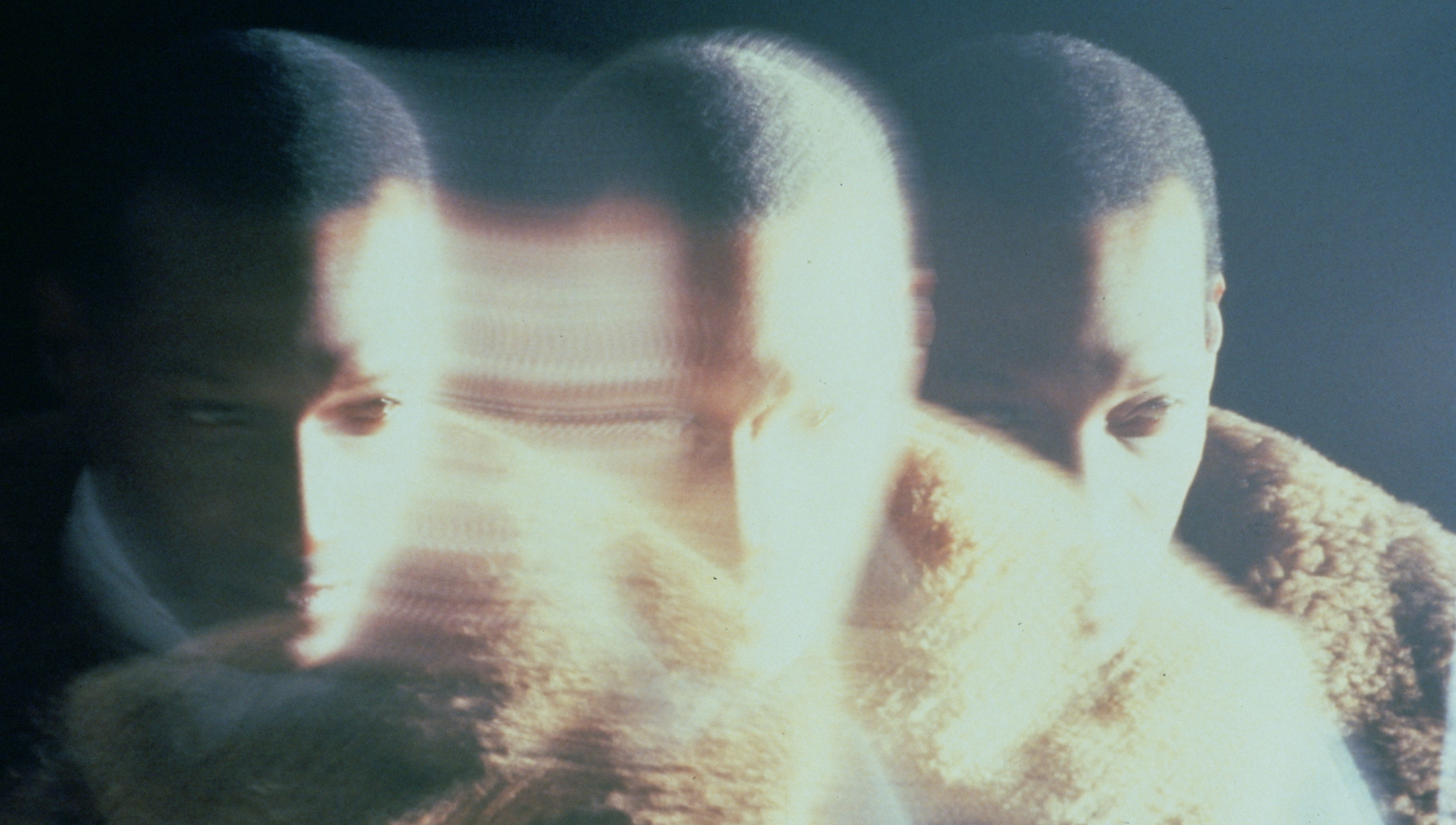 Three blurred, mirrored images of a man wearing a fur coat from the neck up.