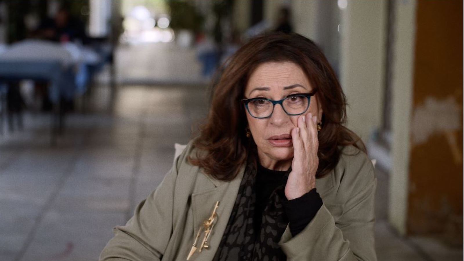 A bespectacled woman in a beige jacker sits in an outdoor restaurant, her hand on her cheek with a befuddled expression.