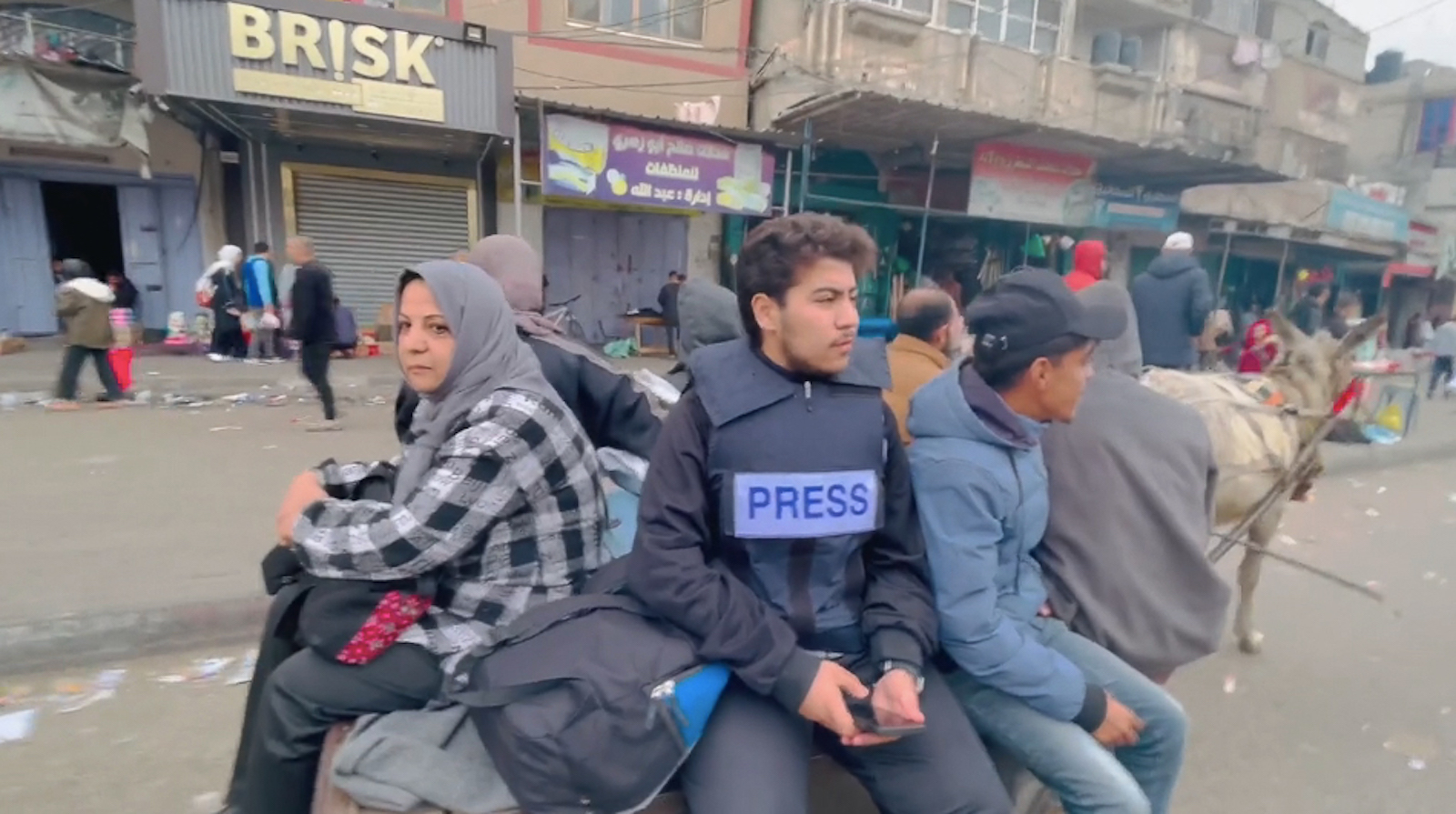 A group of young people sit on the back of a truck in a war-torn middle eastern city; the man in the middle wears a bulletproof vest with the word PRESS on it.