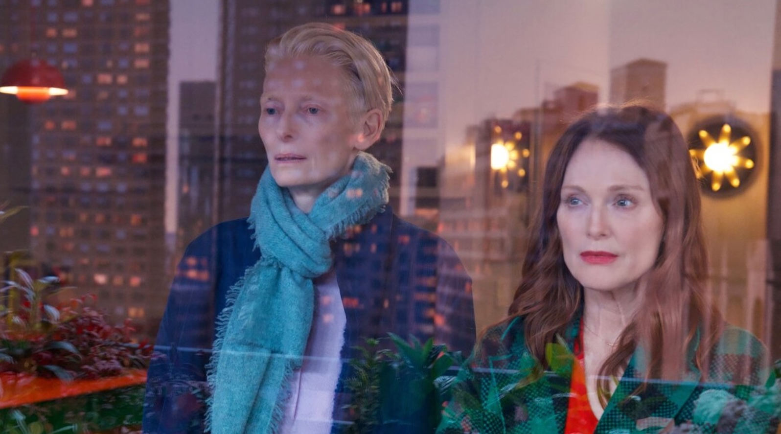 Two women, one with short white hair and wearing a blur scarf, the other with a red shirt and green jacket, stare out a New York City window, with the city skyline reflected on them.