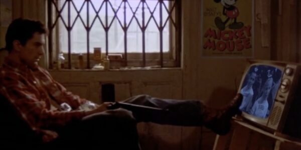 A man sits in a dark afternoon apartment with one foot on a small TV as though he's pushing it over to break it.