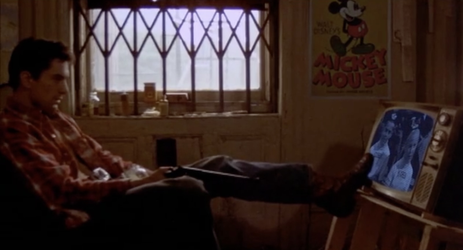 A man sits in a dark afternoon apartment with one foot on a small TV as though he's pushing it over to break it.