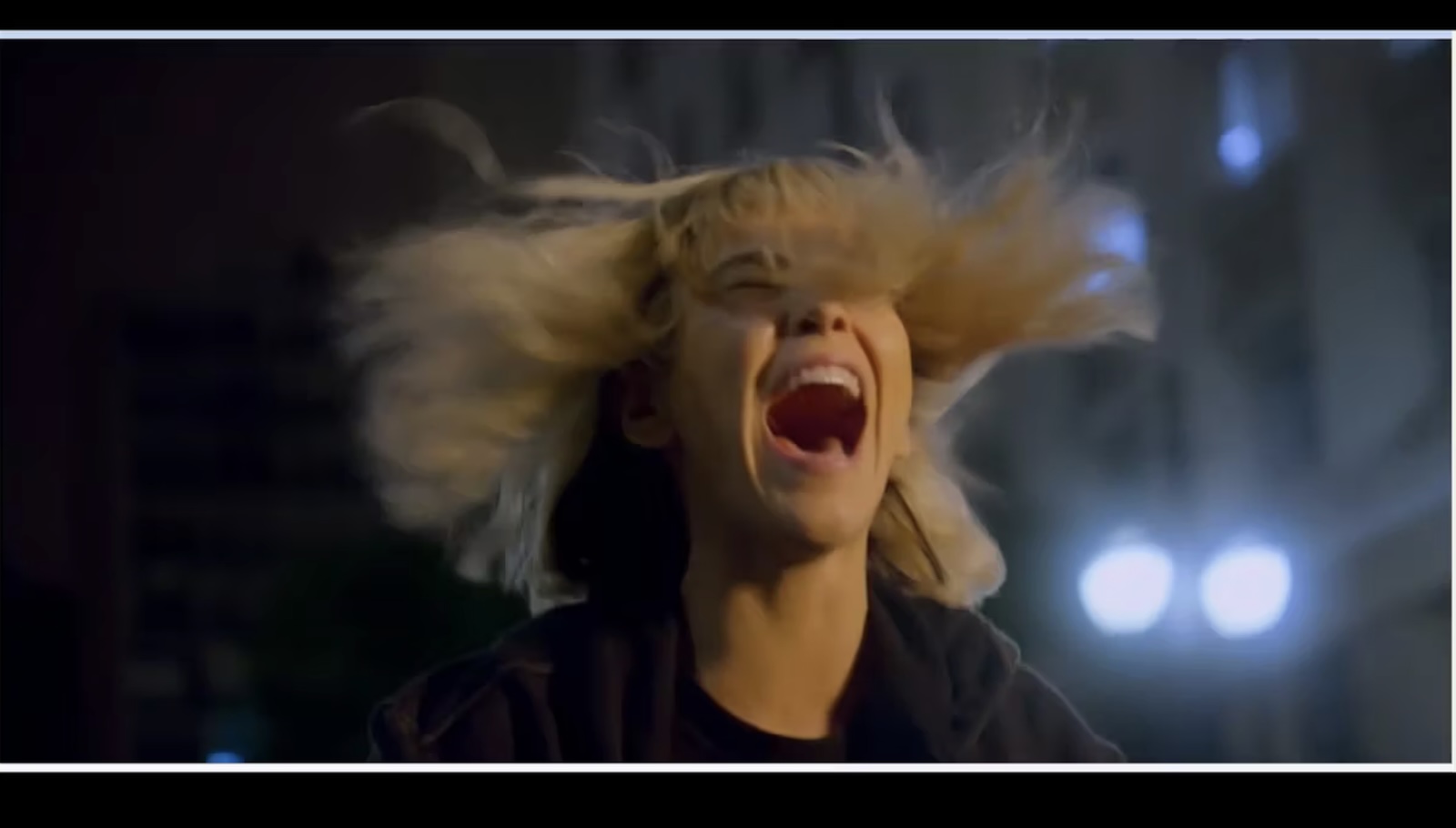 A woman with tousled blonde hair blowing in the wind screams with her mouth wide open.