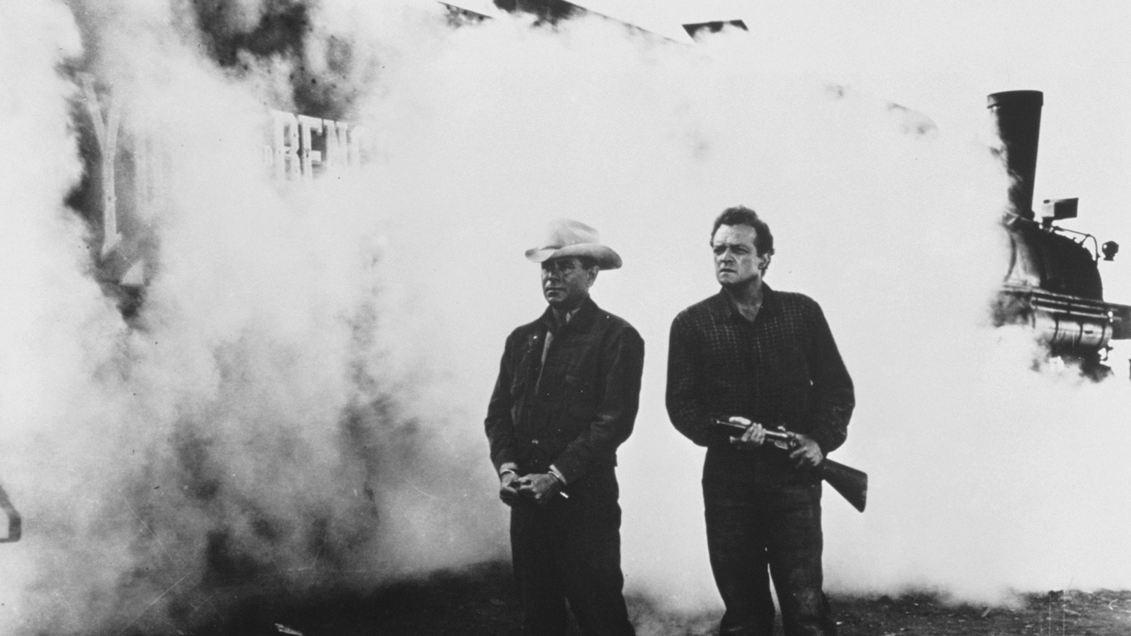 Two men in old west clothes stand against a train surrounded by thick engine steam; one wears a stetson hat and is handcuffed, the other holds a rifle