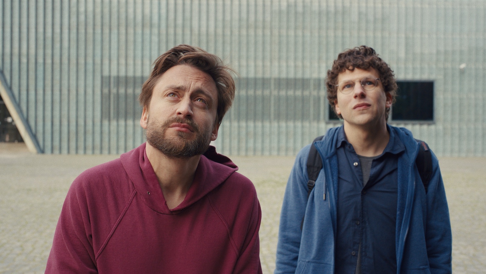 Two young men, one wearing red and the other blue, look up to the sky with bemused painful expressions.