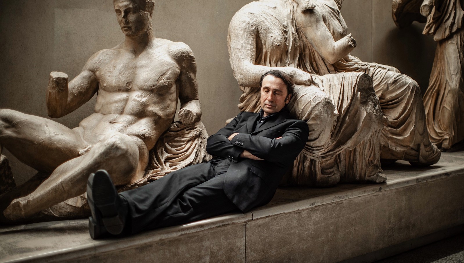 A man in a black suit leans back against classical Greek statues, asleep.