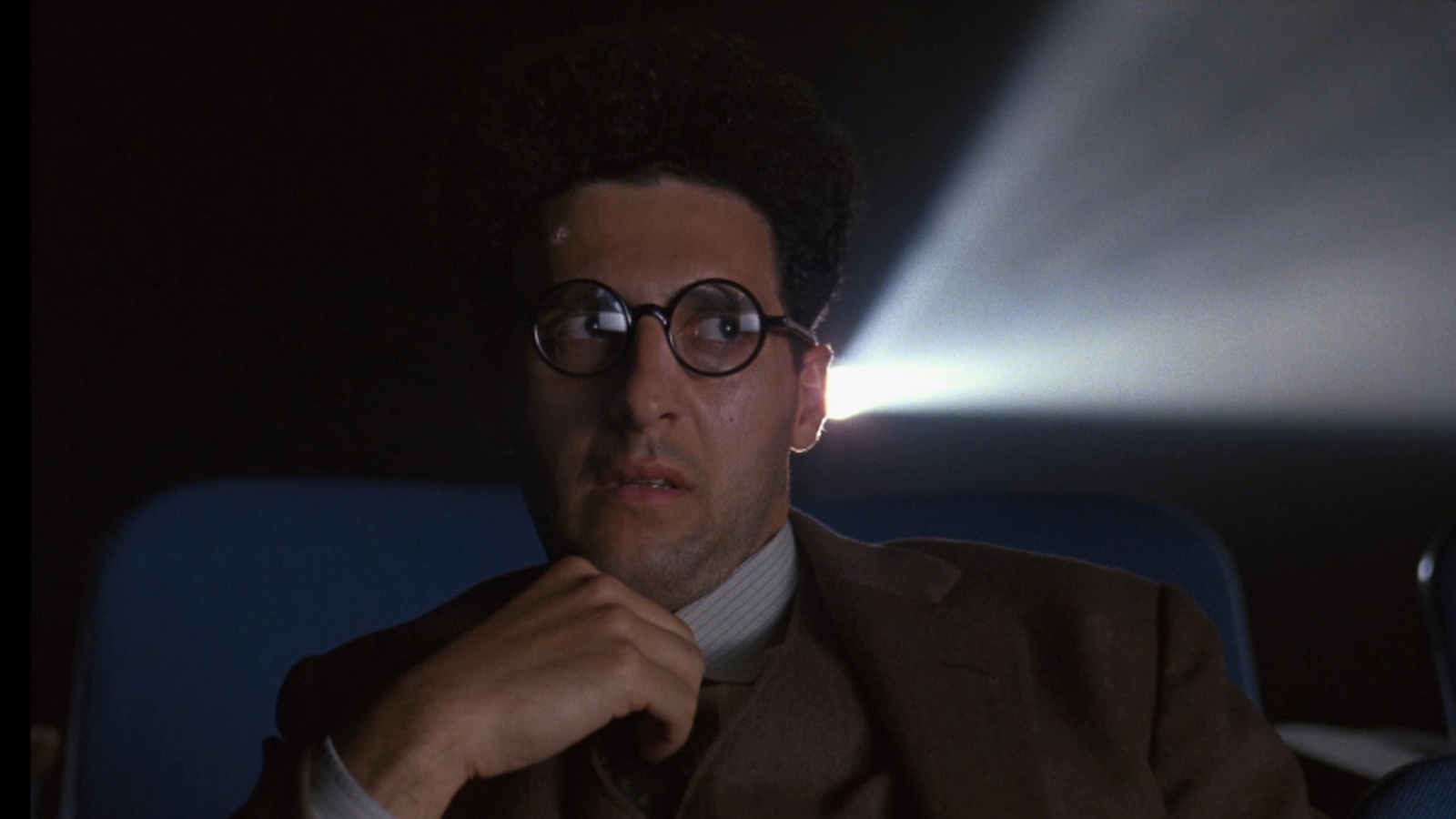A bespectacled man looks aghast as he sits in a movie theater with a projector light cast behind him.