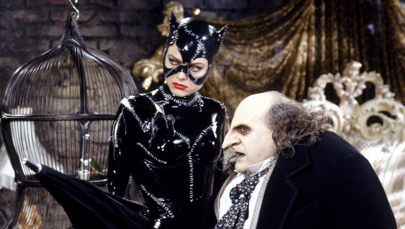A woman dressed in a shiny leather catsuit stitched together with thread and a catmask covering most of her face, stands next to a much shorter, rotund man with scraggly gray hair and a long nose, holding a closed umbrella.