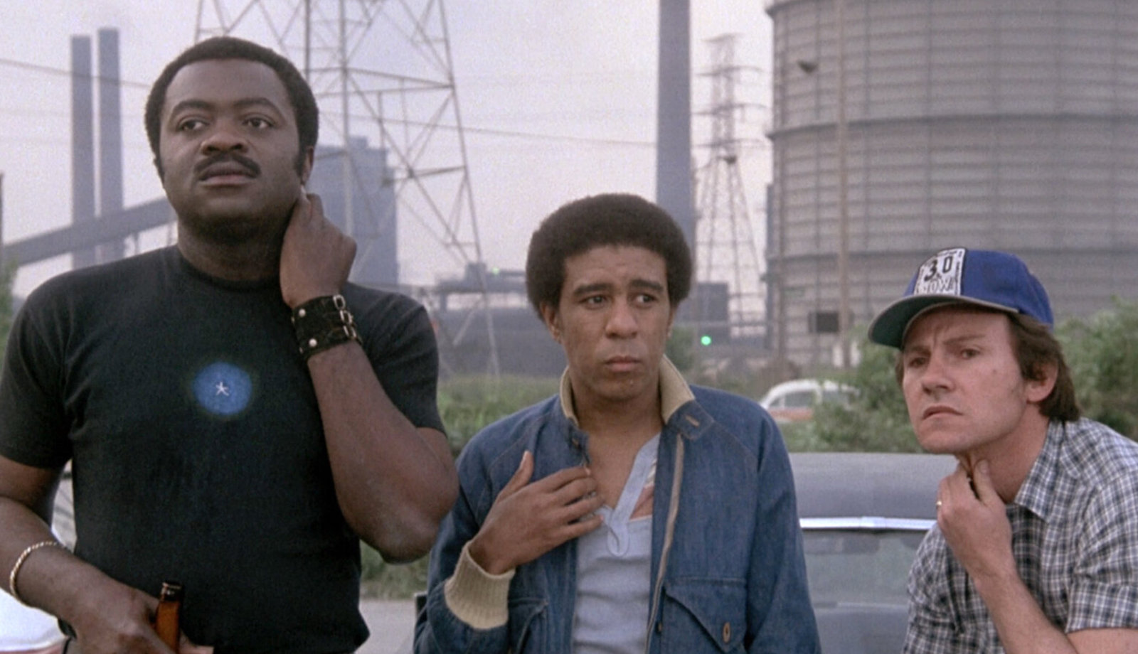 Three men stand against an industrial landscape.