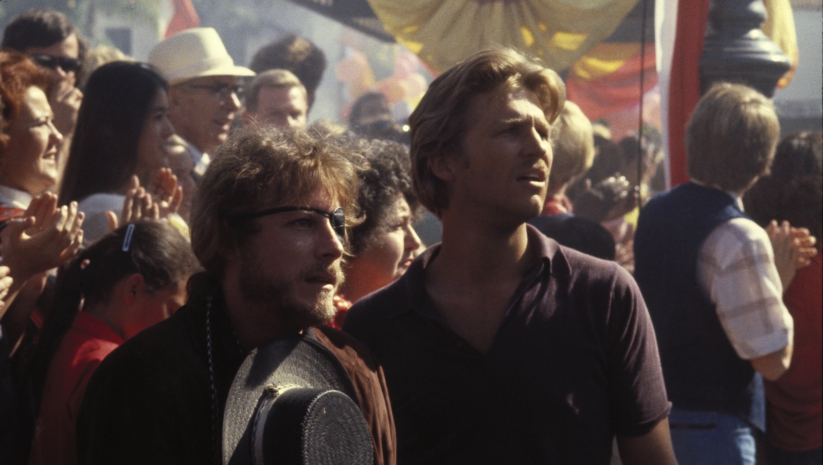 Two men, one with an eye-patch, stand in a crowd, looking off into the distance with unsettled expressions.