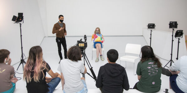 Two face-masked people, a standing instructor in a brown cardigan and a sitting young woman in a tie-dyed colorful shirt, appear before a group of students, their backs to the camera, in an all-white room, with klieg light stands on either side.