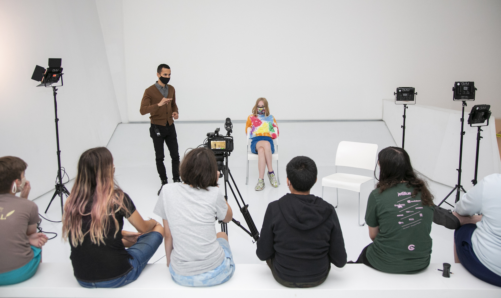 Two face-masked people, a standing instructor in a brown cardigan and a sitting young woman in a tie-dyed colorful shirt, appear before a group of students, their backs to the camera, in an all-white room, with klieg light stands on either side.