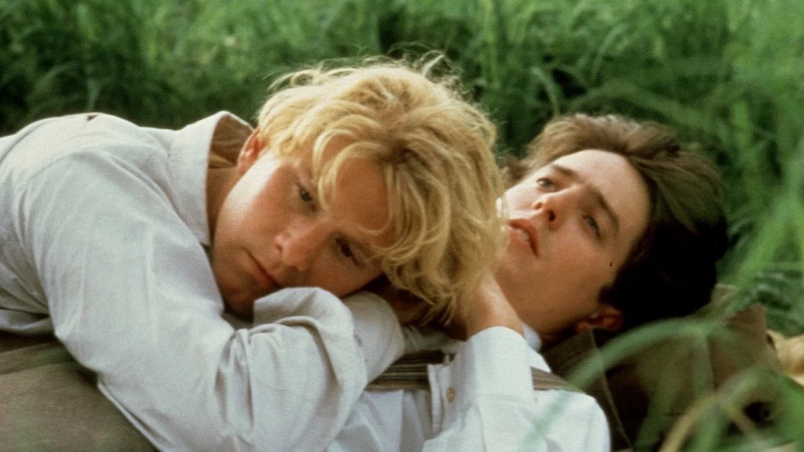 A blond man lies on a dark-haired man's chest in a field of tall green grass, both wearing white dress shirts.