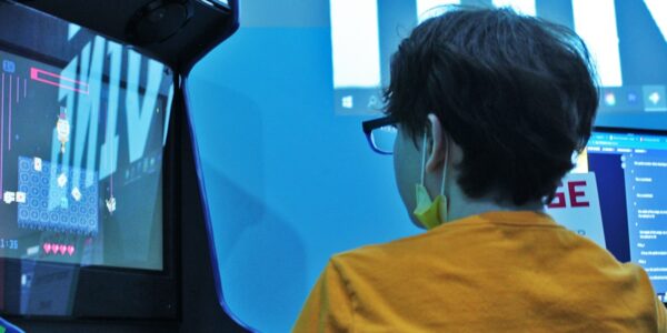 From behind, a boy in a yellow shirt with a face-mask dangling from his chin, plays a video arcade game.