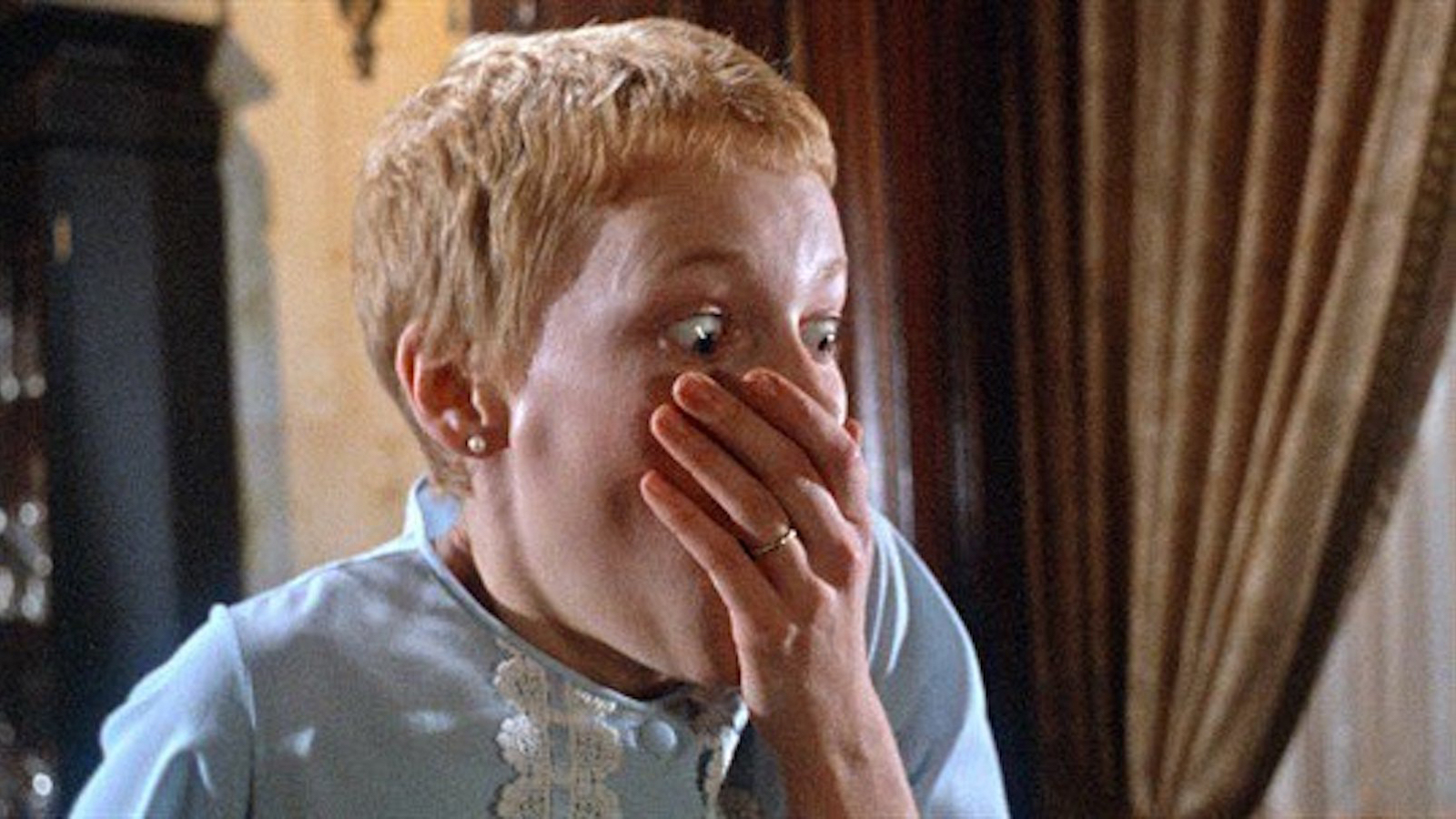 A woman with short hair and a sky-blue nightgown covers her mouth in horror.