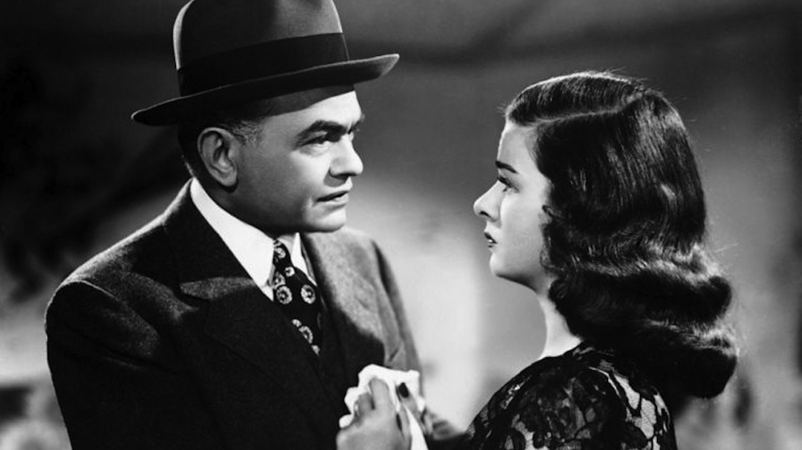 A man in a derby hat and suit stares at a woman with long dark hair holding a handkerchief with a serious expression.