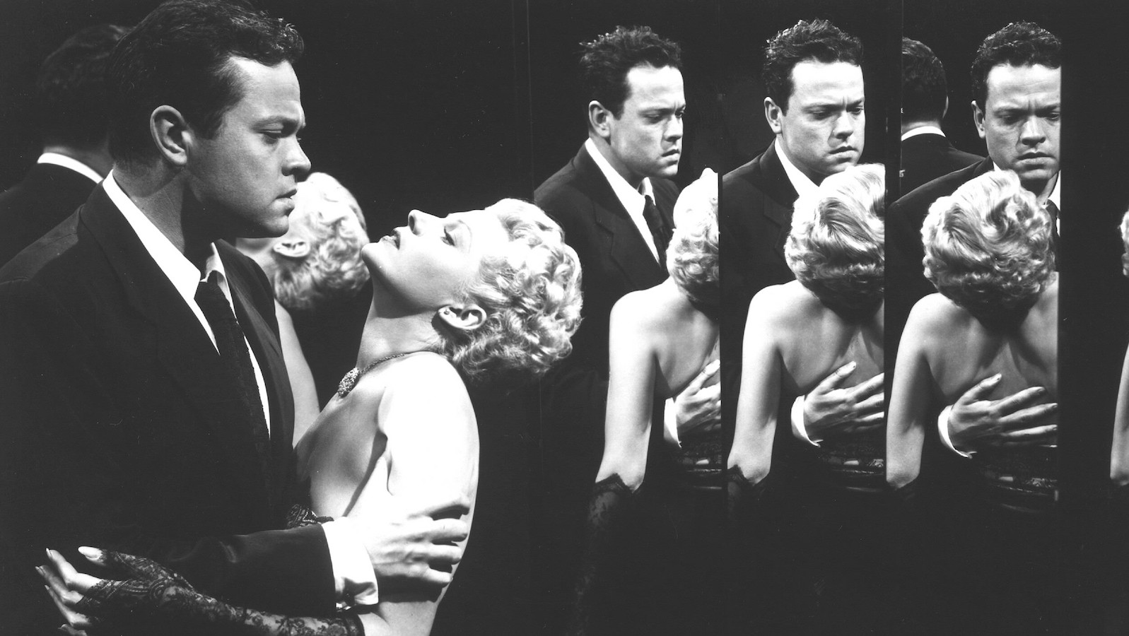 A suited man clutches a short-haired blonde woman, her head back 90 degrees, in a dramatic pose as they stand against a series of mirrors that creates a hall of reflections.