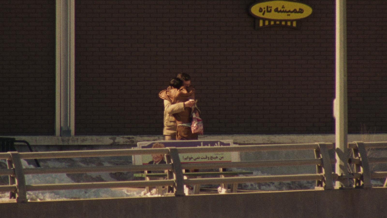 Two people hug on a city street from a distance, one clutching a bag of unidentifiable food. There is Farsi writing against the building behind them.