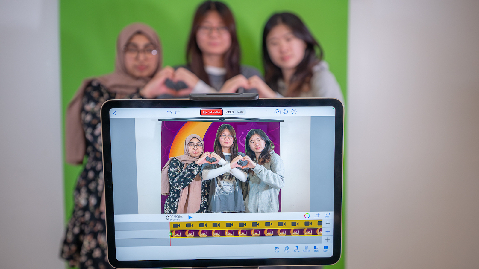 Three young women pose in front of a camera, captured on a computer screen in the foreground, holding their hands together to make heart shapes.